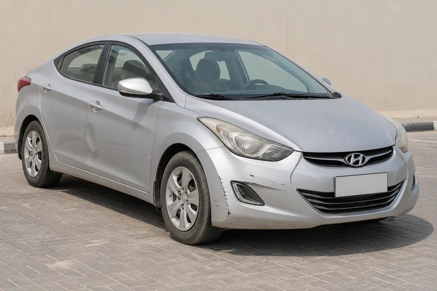 Used early-2010s Elantra-style Hyundai sedan with front bumper scuffs and a shallow door dent at a UAE assessment forecourt