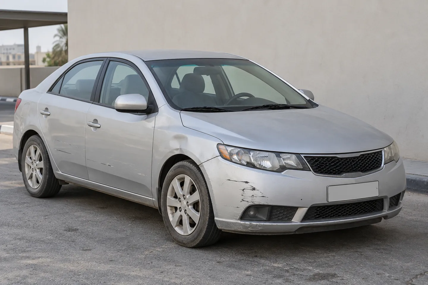 Used early-2010s Cerato-style Kia sedan with front bumper scrapes, a shallow fender dent and door scratches at a UAE assessment forecourt