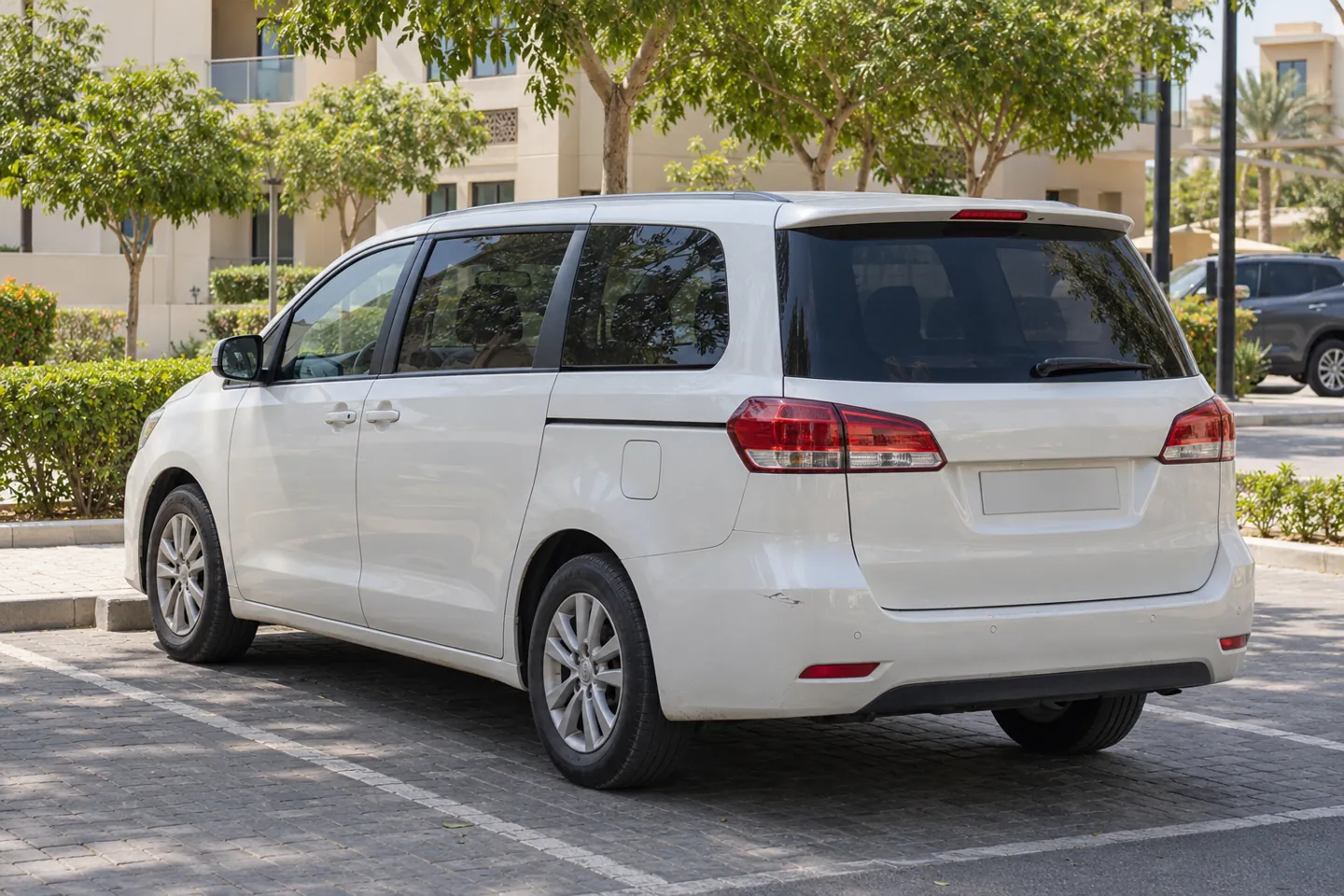 Pearl-white mid-2010s family MPV with modest wear viewed from the rear in an Abu Dhabi community outdoor parking bay beside apartment buildings and landscaping