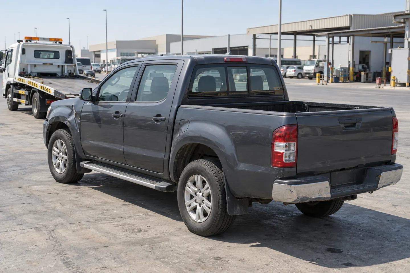 Dark-grey mid-2010s double-cab pickup with modest fender damage viewed from the rear beside an unbranded flatbed in a Musaffah-style industrial service area